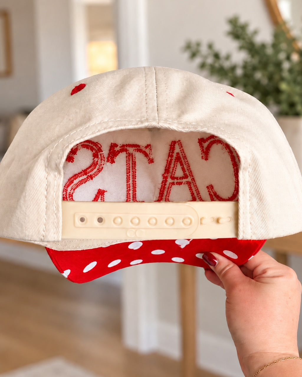 CATS Pep Rally Hat held from the back to show the cream snapback closure and red polka-dot bill.