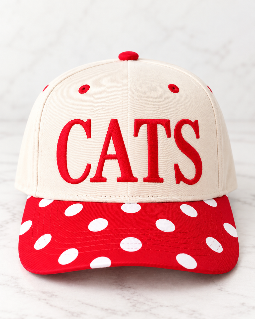 Front view of cream CATS Pep Rally Hat with red embroidered lettering and a red bill with white polka dots.
