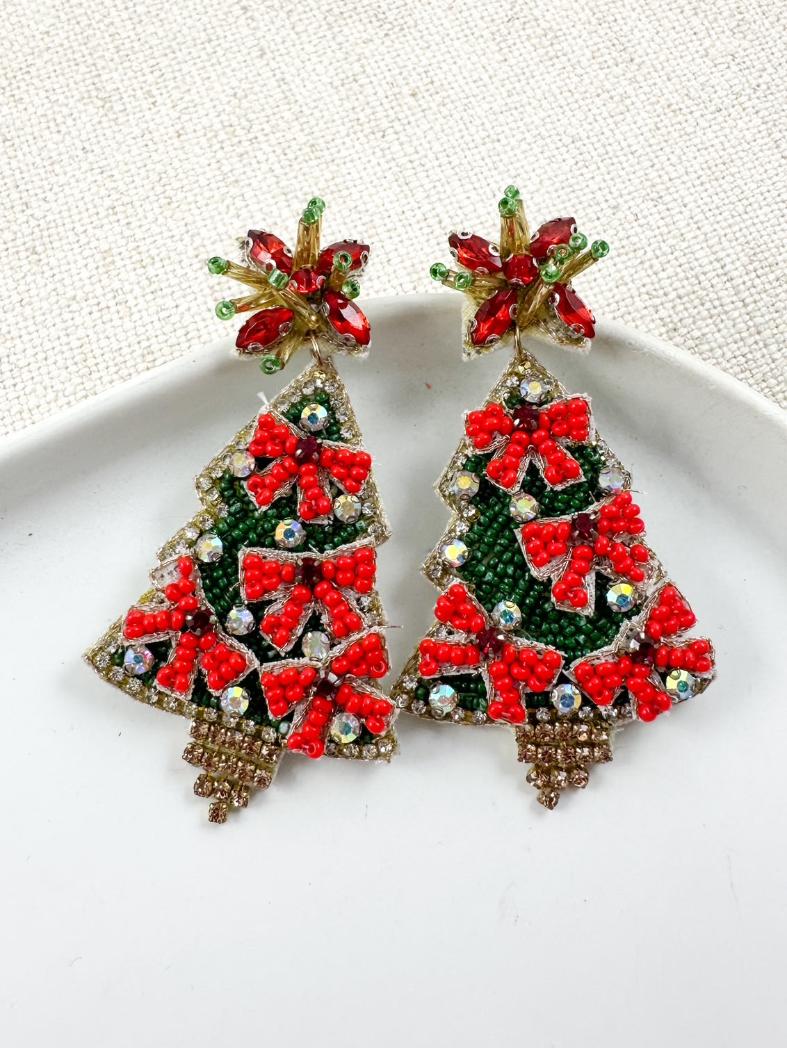 Beaded Christmas tree earrings with green beadwork, red bow accents, crystal details, and jeweled star studs displayed on a white dish.