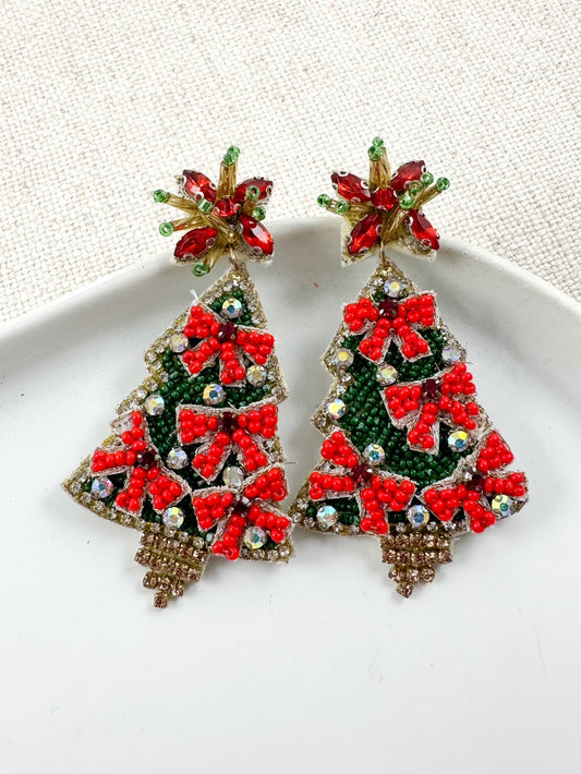 Beaded Christmas tree earrings with green beadwork, red bow accents, crystal details, and jeweled star studs displayed on a white dish.