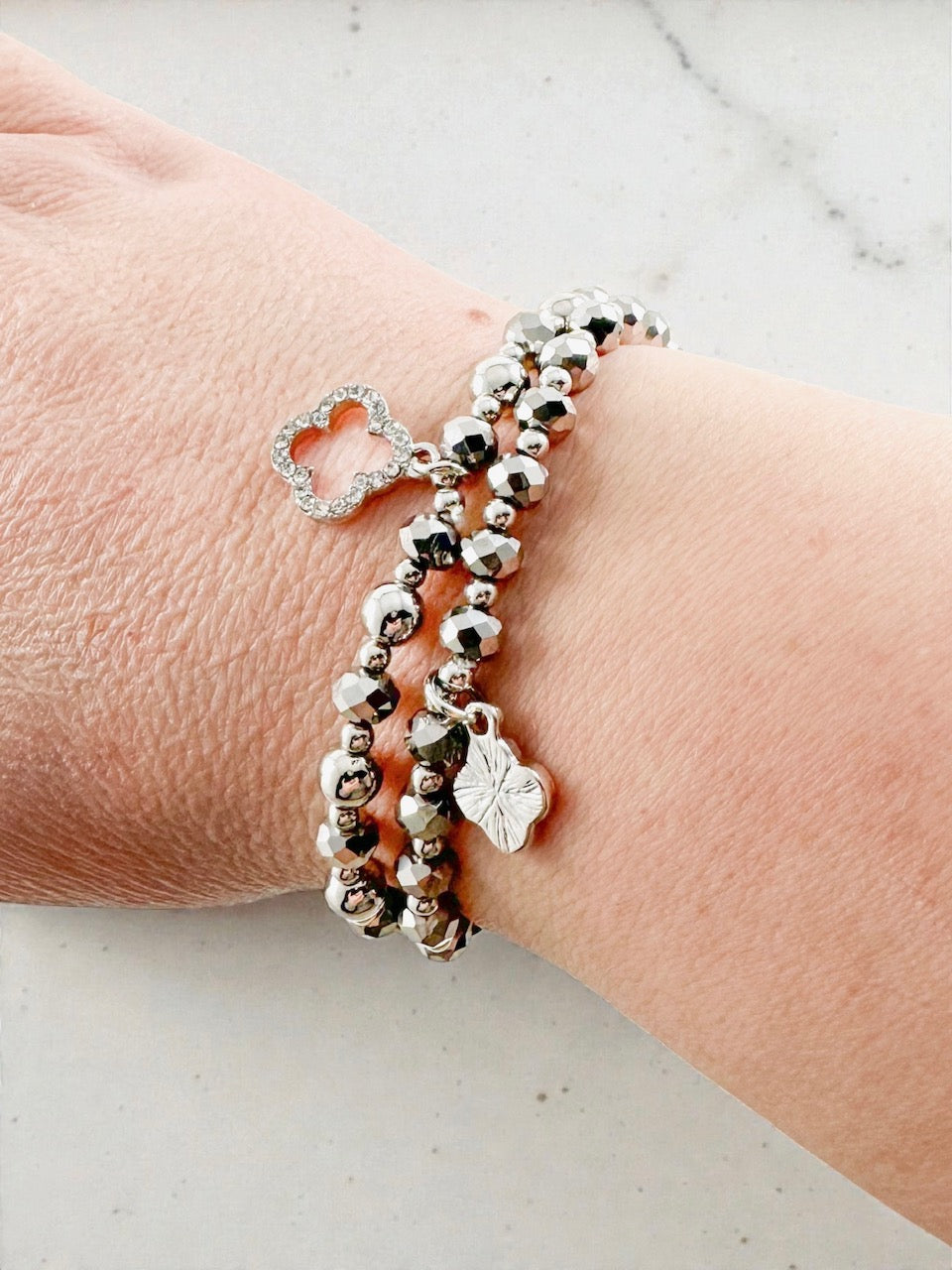 Silver beaded stretch bracelet set worn on a wrist, featuring faceted beads and a clover charm.