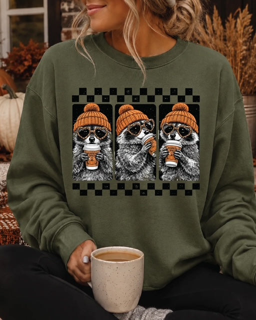Woman wearing the green Cozy Little Bandits sweatshirt featuring three raccoons in orange beanies sipping coffee.