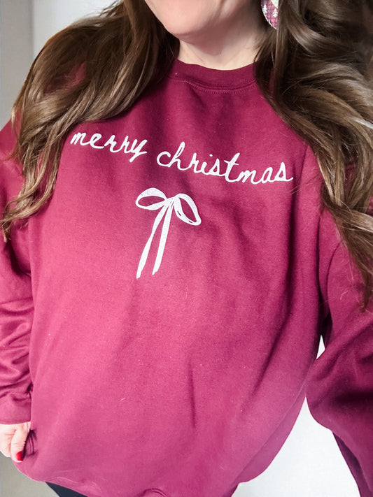 Woman wearing a cranberry Merry Christmas sweatshirt with a small white bow printed underneath the text.