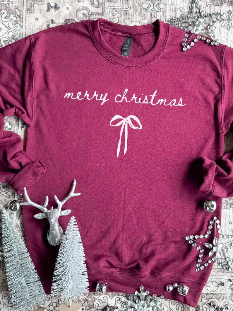 Flat lay of a cranberry Merry Christmas sweatshirt with a small white bow, surrounded by silver Christmas decor including trees, a reindeer, and beads.