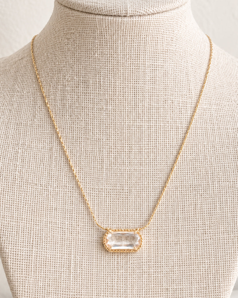 Crystal Clear Necklace displayed on a cream linen jewelry bust, showing how the clear rectangular pendant hangs from the delicate gold-tone chain.