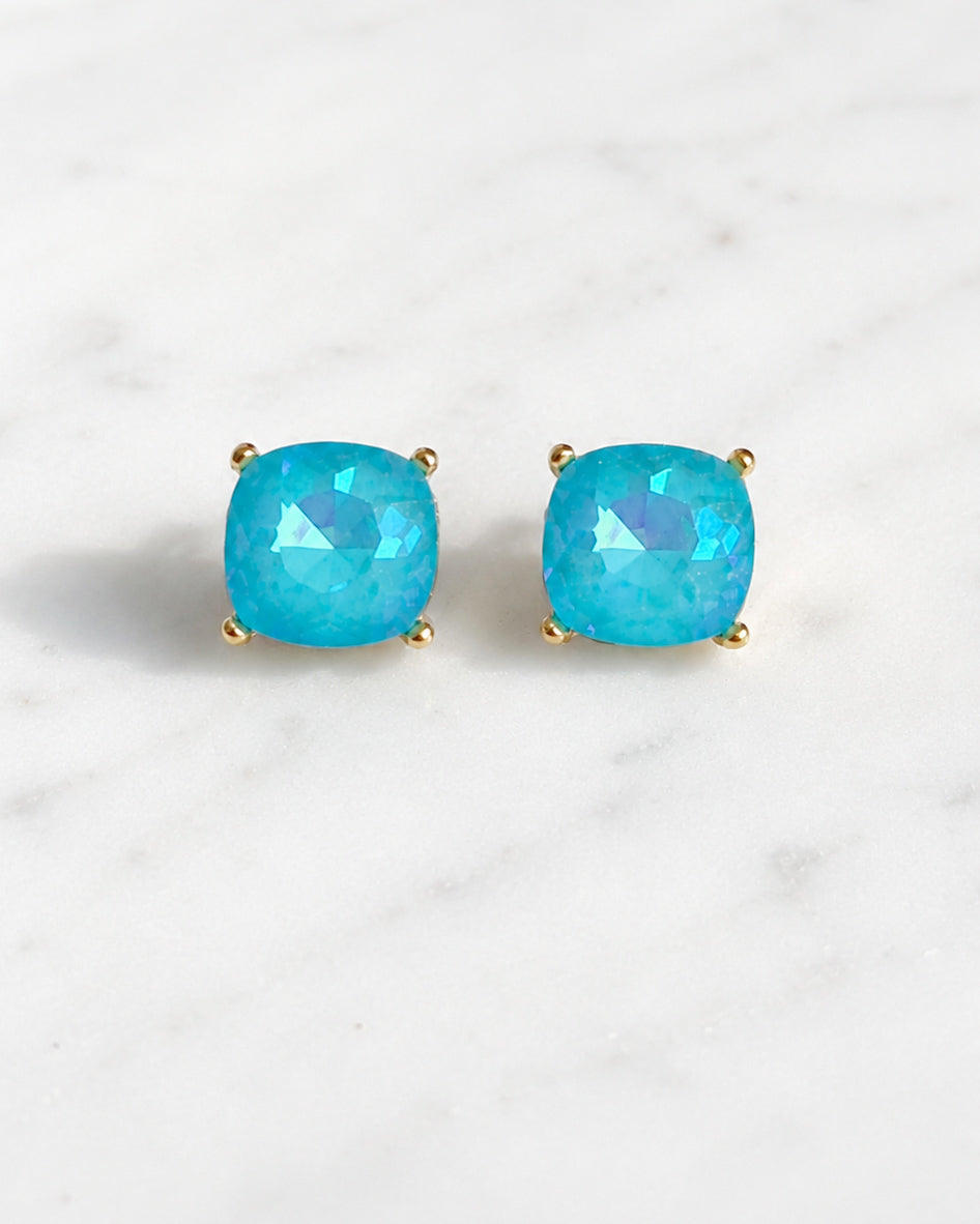 Close-up of turquoise iridescent square stud earrings with gold-tone hardware on a white marble background.
