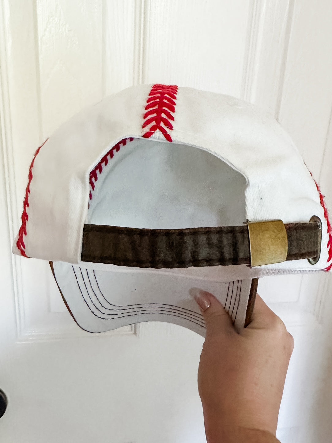 Back of white baseball cap with red embroidered stitching and adjustable brown strap with metal clasp.