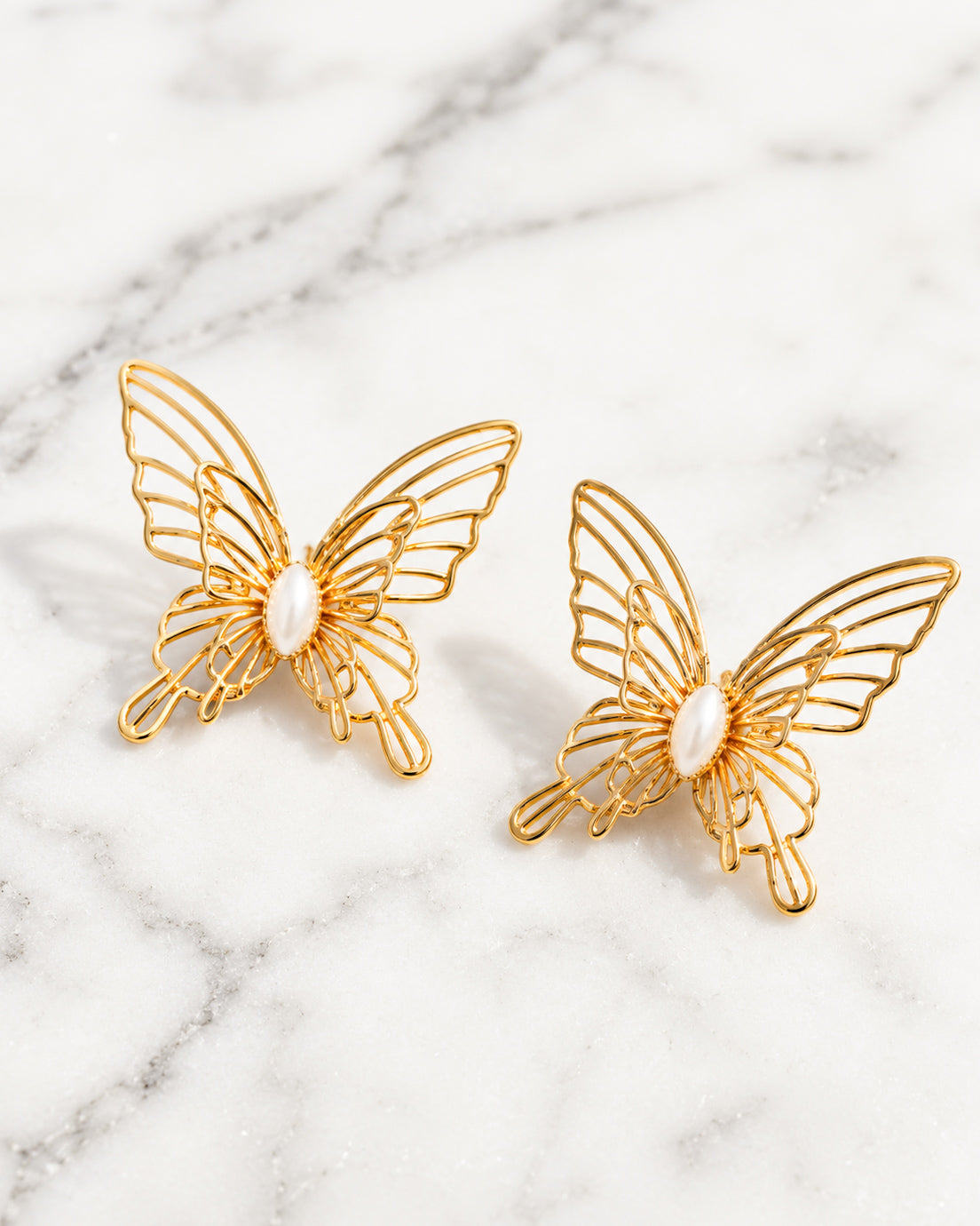 Gold butterfly stud earrings with intricate filigree details and faux pearl centers on white marble.