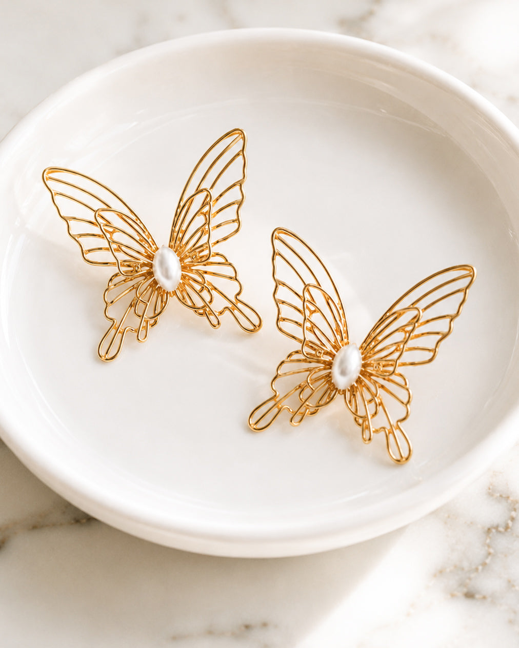 Gold filigree butterfly stud earrings with faux pearl centers resting in a white ceramic jewelry dish.