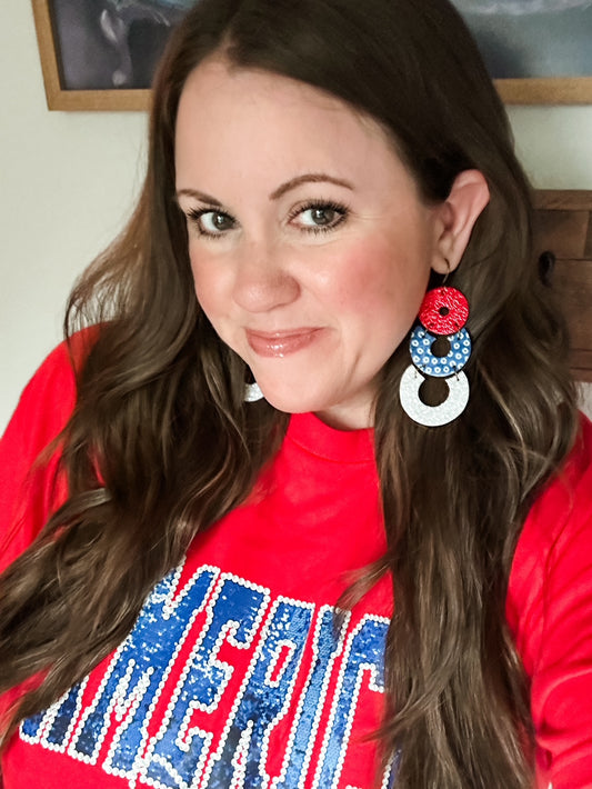 Firecracker Pop Earrings modeled in the large size featuring red sparkle, blue dotted cork, and white shimmer genuine leather details.