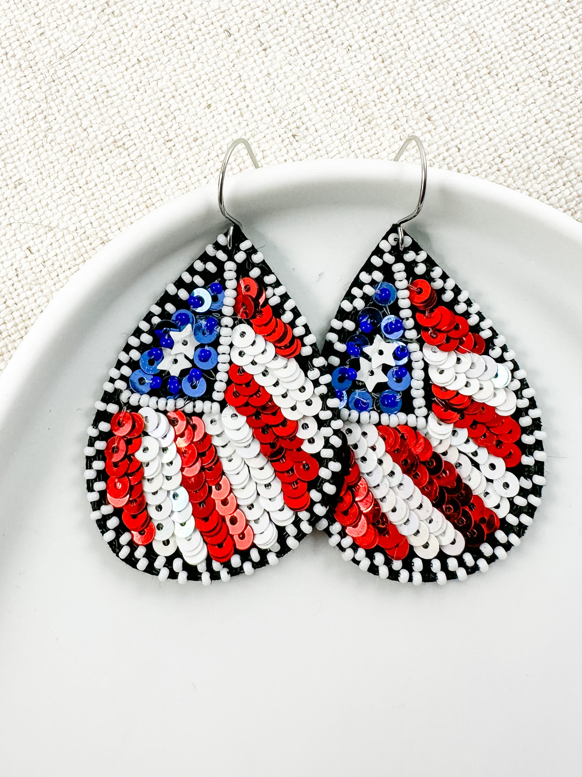 Flag of Freedom patriotic statement earrings featuring red, white, and blue sequin American flag details with black and white bead accents displayed in a white ceramic dish.
