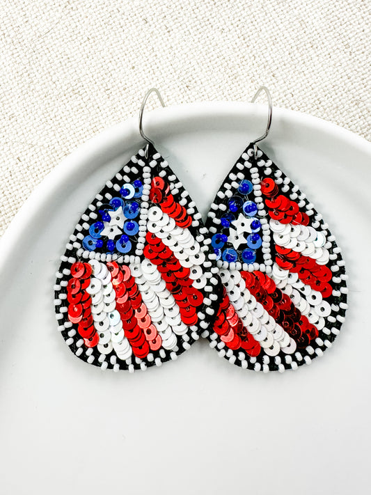 Flag of Freedom patriotic statement earrings featuring red, white, and blue sequin American flag details with black and white bead accents displayed in a white ceramic dish.