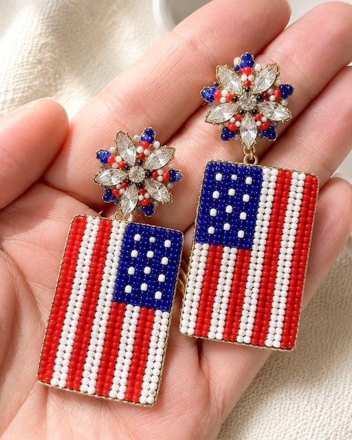 Freedom Rings patriotic seed bead earrings featuring American flag drops and sparkling red, white, and blue starburst studs shown in hand for scale.