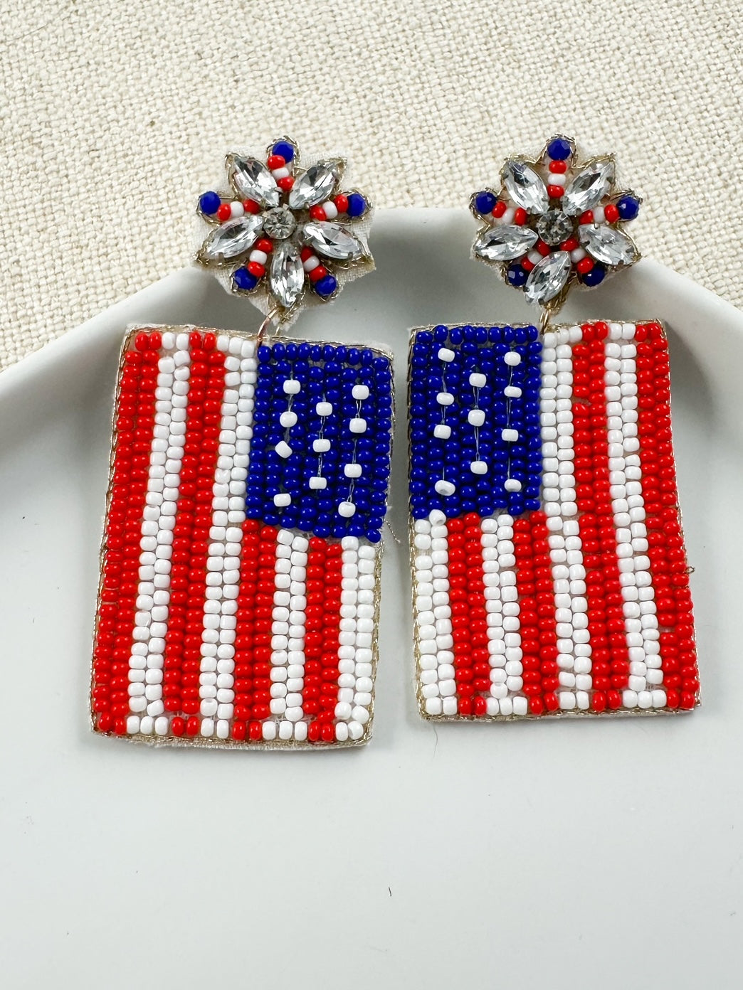 Freedom Rings patriotic statement earrings with seed bead American flag drops and crystal starburst stud tops displayed on a white dish.