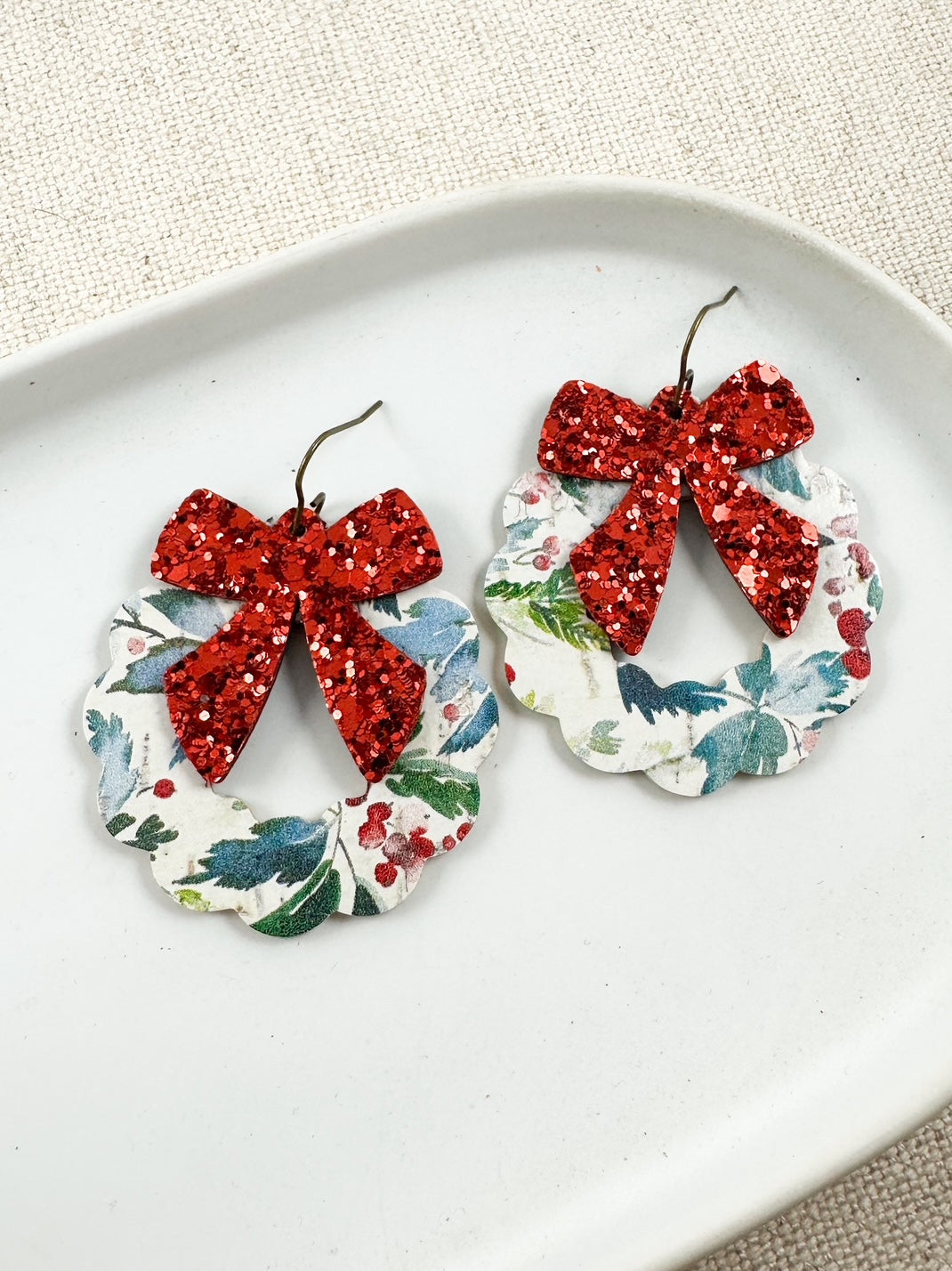 Frosted Berry Wreath Earrings with red glitter bows and floral cork wreaths displayed on a white dish.