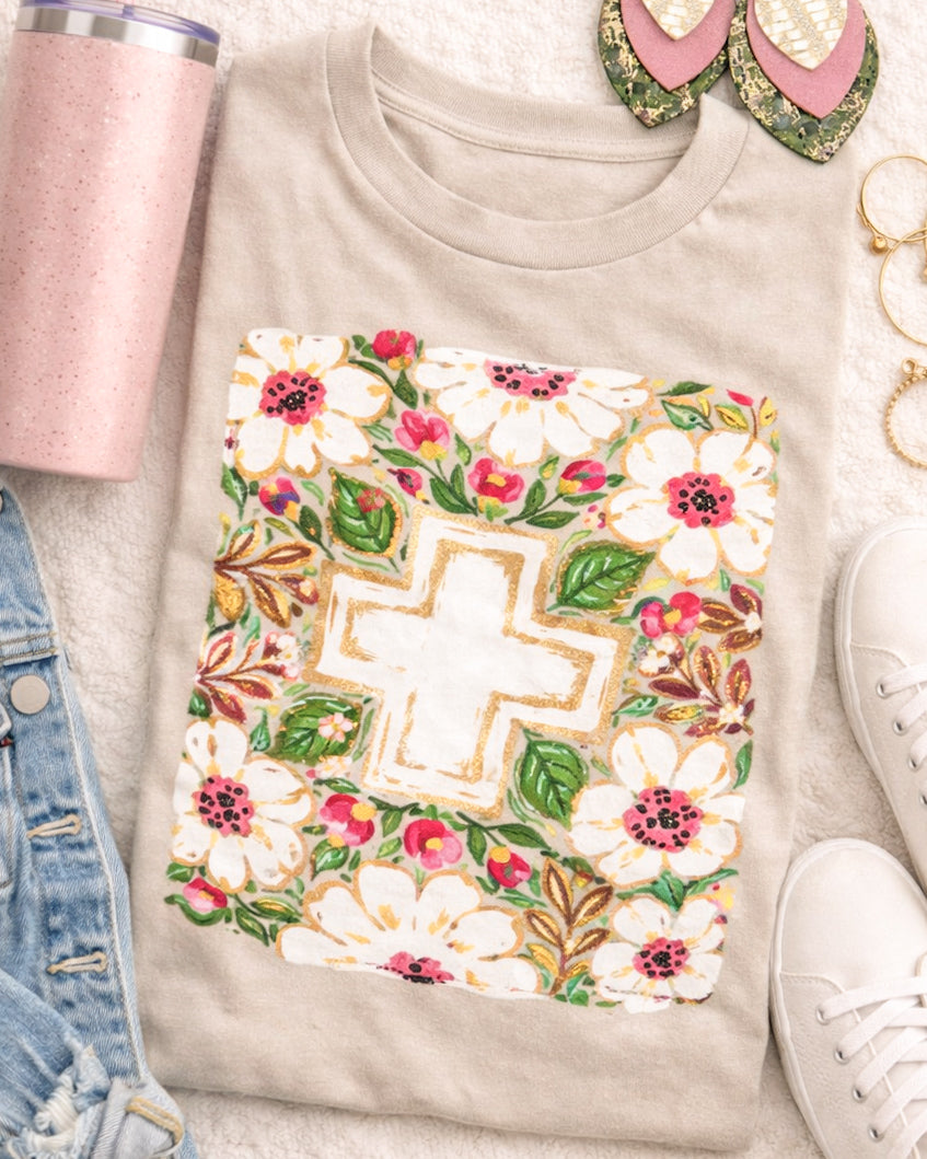 Heather tan Bella Canvas Christian graphic tee with floral cross design styled in a flat lay with denim jacket, bracelets, sneakers, and a pink tumbler.