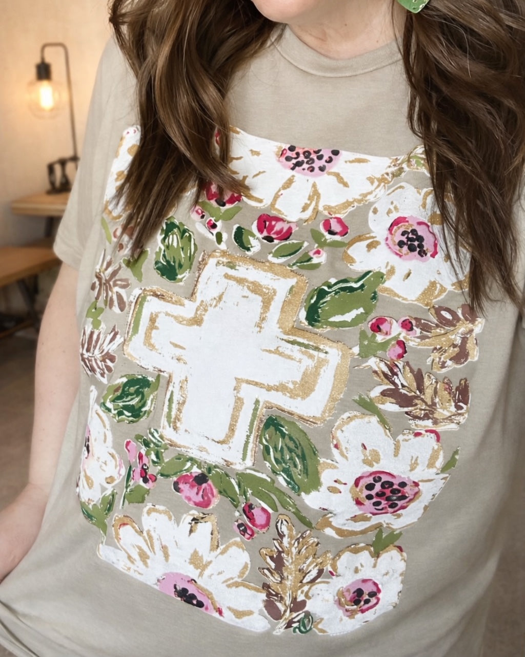 Woman wearing a heather tan Christian graphic tee featuring a floral cross design with pink flowers, green leaves, and gold accents.