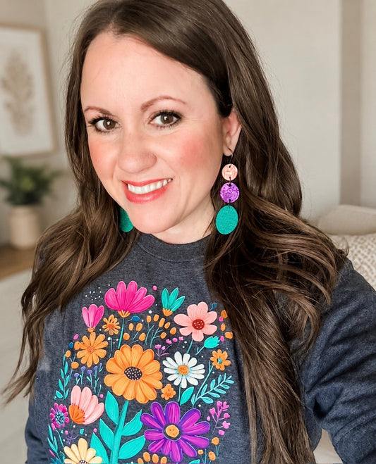 Woman wearing handmade Garden Party leather and cork dangle earrings with coral, purple, and emerald green circles paired with a colorful floral graphic tee.