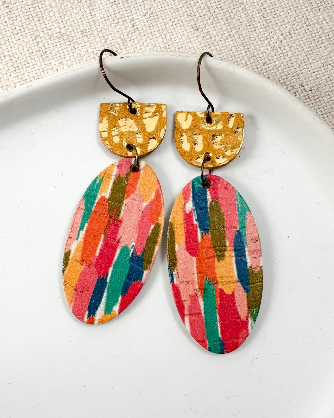 Close-up of Gilded Brushstrokes leather and cork earrings featuring textured gold semicircle tops and multicolored oval drops in a white ceramic dish.