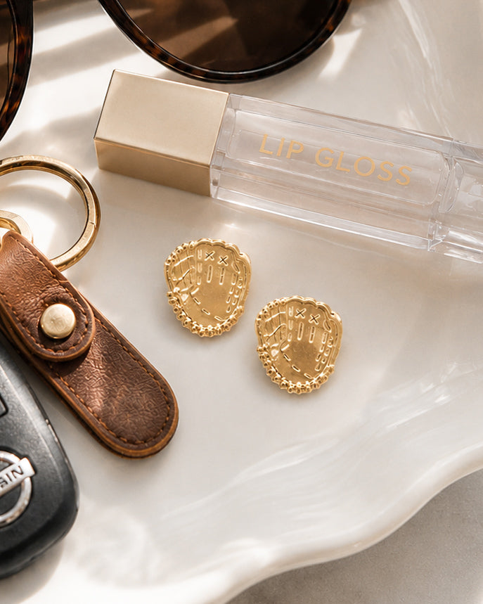 Gold baseball glove stud earrings on white marble with everyday accessories including sunglasses, lip gloss, and keys.