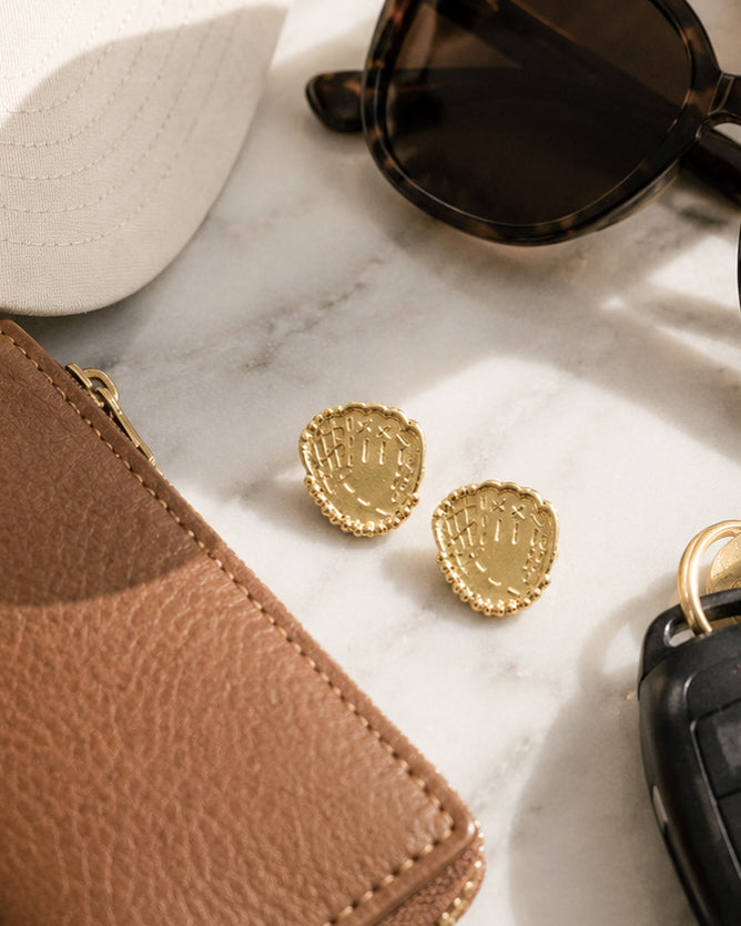 Gold baseball glove stud earrings styled with a baseball cap, wallet, sunglasses, and keys on marble.