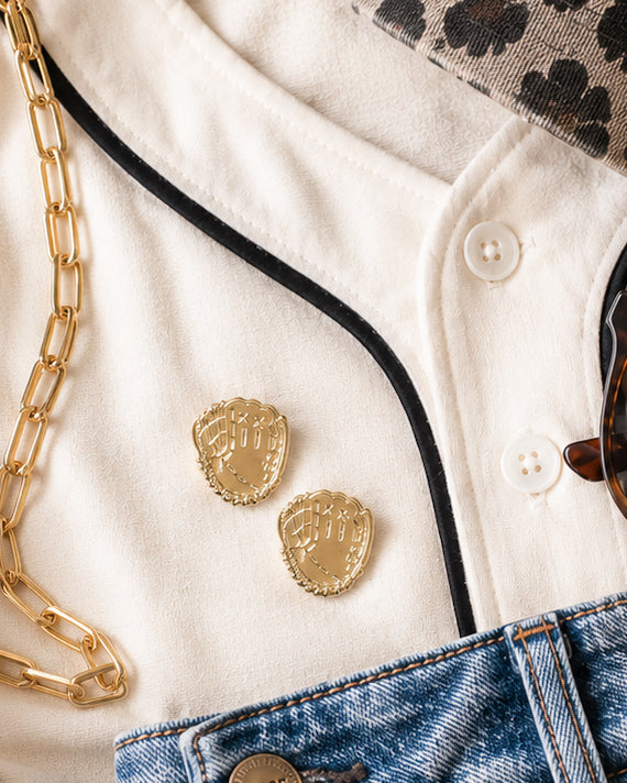 Gold baseball glove stud earrings styled with a cream jersey, denim jeans, and gold accessories for a casual baseball or softball game day outfit.