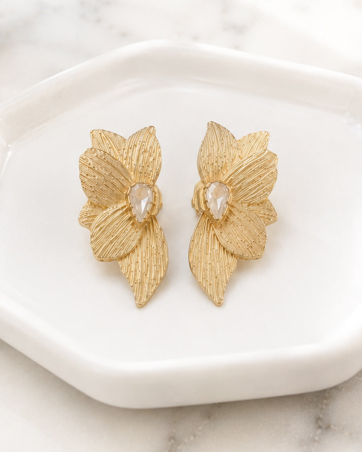 Gold floral statement earrings resting in a white ceramic dish with soft natural lighting.