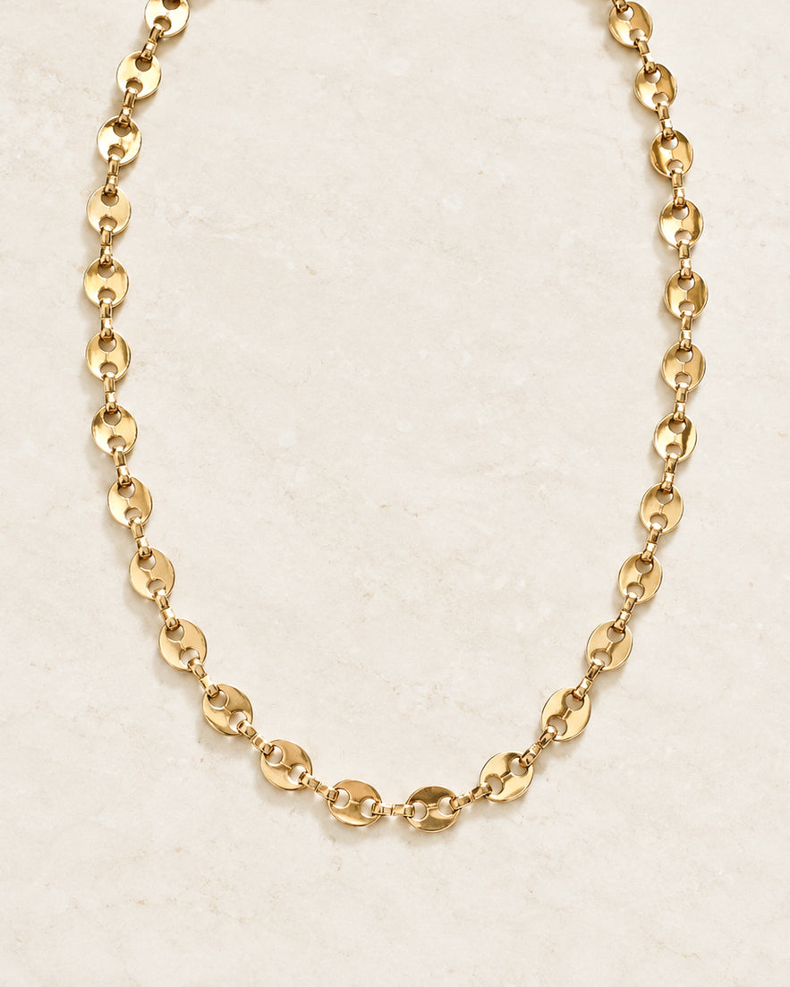 Close-up of the Golden Together Necklace showing shiny gold-tone oval links connected by smaller chain links, arranged on a light neutral stone surface.