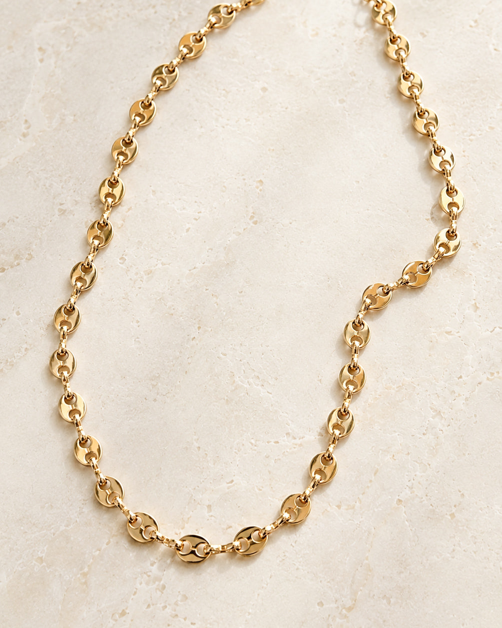 Golden Together Necklace featuring polished gold-tone oval pig nose chain links arranged naturally on a textured cream stone background.