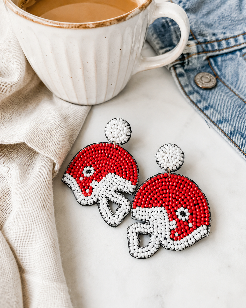 Red football helmet seed bead statement earrings on white marble beside a cream coffee mug, ivory linen, and denim jacket.