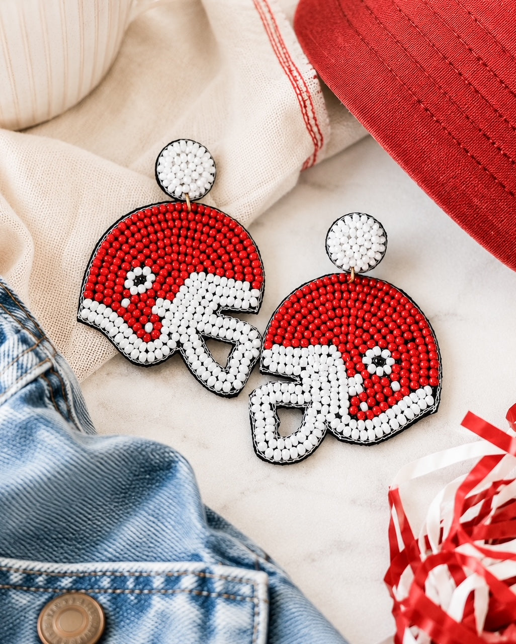 Red football helmet seed bead earrings styled with a red baseball cap, denim jacket, and ivory linen for a game day outfit.