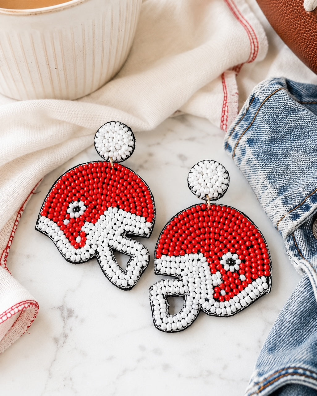 Red football helmet seed bead statement earrings on white marble with a football, cream coffee mug, ivory linen, and denim jacket.