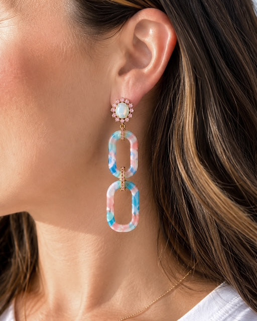 Woman wearing Hampton Drop Earrings featuring blue and pink marbled acrylic links with iridescent opal-like crystal studs and gold accents.