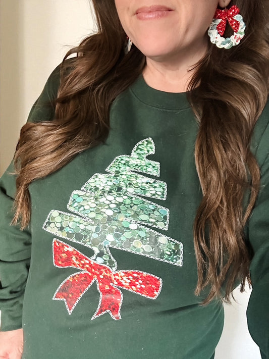 Woman wearing the Holiday Glow Sweatshirt with an evergreen faux sequin tree and red bow graphic.