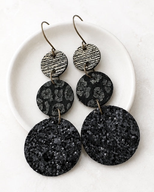 Black leather circle earrings arranged on a white dish with coordinating black and metallic textures