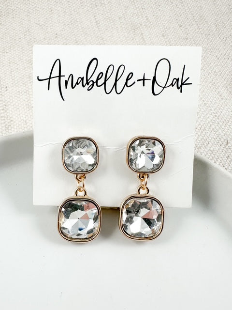 Clear faceted jewel drop earrings with gold settings displayed on an Anabelle+Oak card on a white plate.