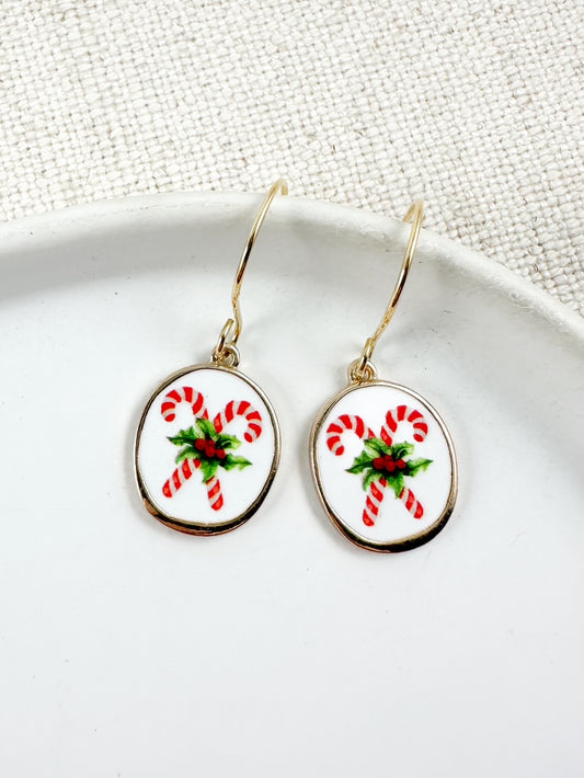 Oval drop earrings with white background and red candy cane design accented with green details hanging from gold hooks on a white dish