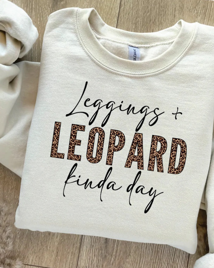 Cream graphic shirt folded flat with black script lettering and bold leopard print block text reading “Leggings + Leopard kinda day.”