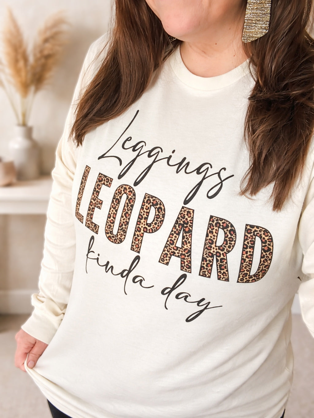 Woman wearing cream long sleeve graphic tee with black script and leopard block lettering reading “Leggings + Leopard kinda day.”