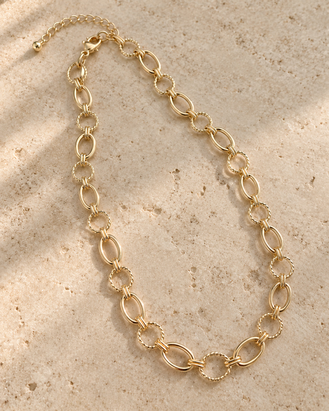 Linked & Lovely Necklace arranged on beige stone, showing alternating polished gold oval links, textured circular links, and an adjustable lobster clasp closure.