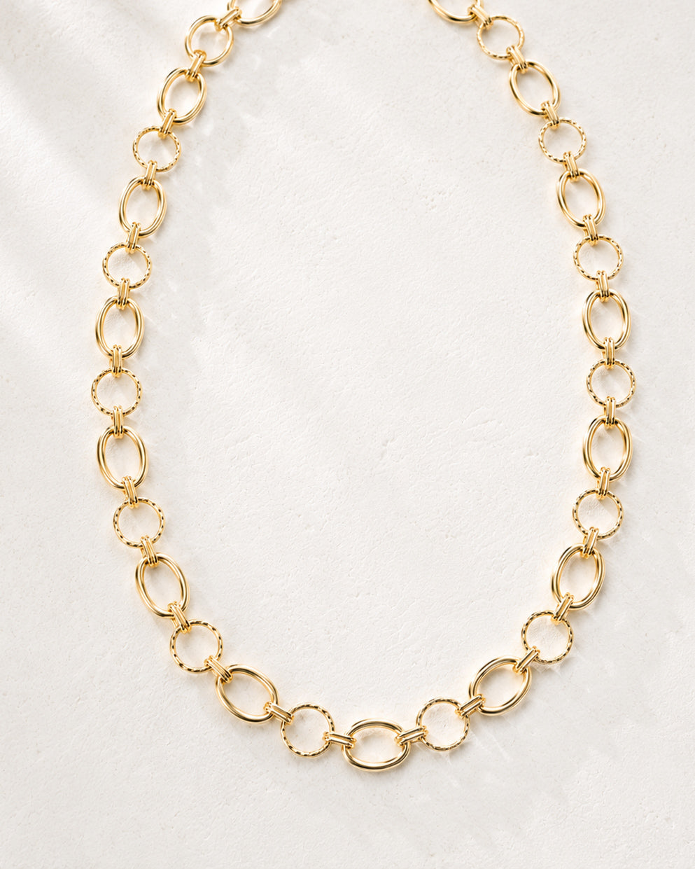 Linked & Lovely gold-tone statement necklace laid in a curved shape on a soft ivory background, highlighting its alternating oval and textured round links.