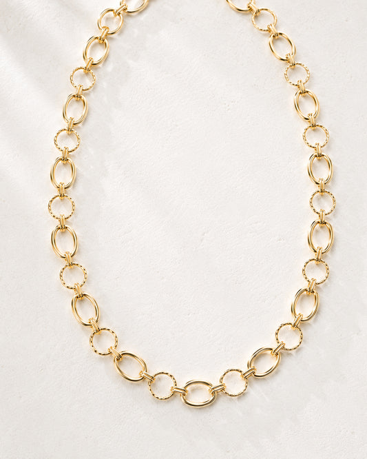 Linked & Lovely gold-tone statement necklace laid in a curved shape on a soft ivory background, highlighting its alternating oval and textured round links.