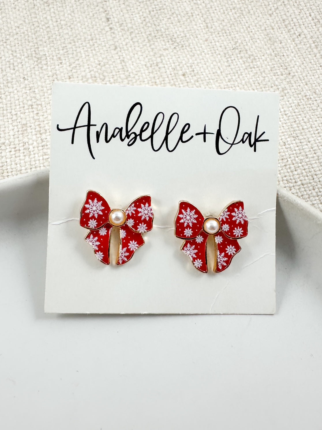 Red bow stud earrings with white snowflake detail and a small pearl center displayed on an Anabelle + Oak branded card.