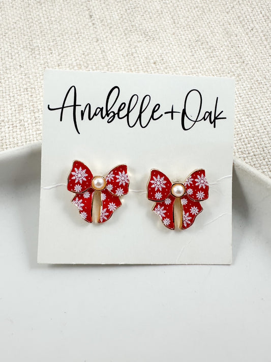Red bow stud earrings with white snowflake detail and a small pearl center displayed on an Anabelle + Oak branded card.