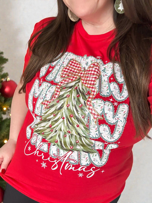 Woman wearing a red Christmas graphic tee with a green quilted-style tree and red gingham bow, standing beside a decorated Christmas tree.