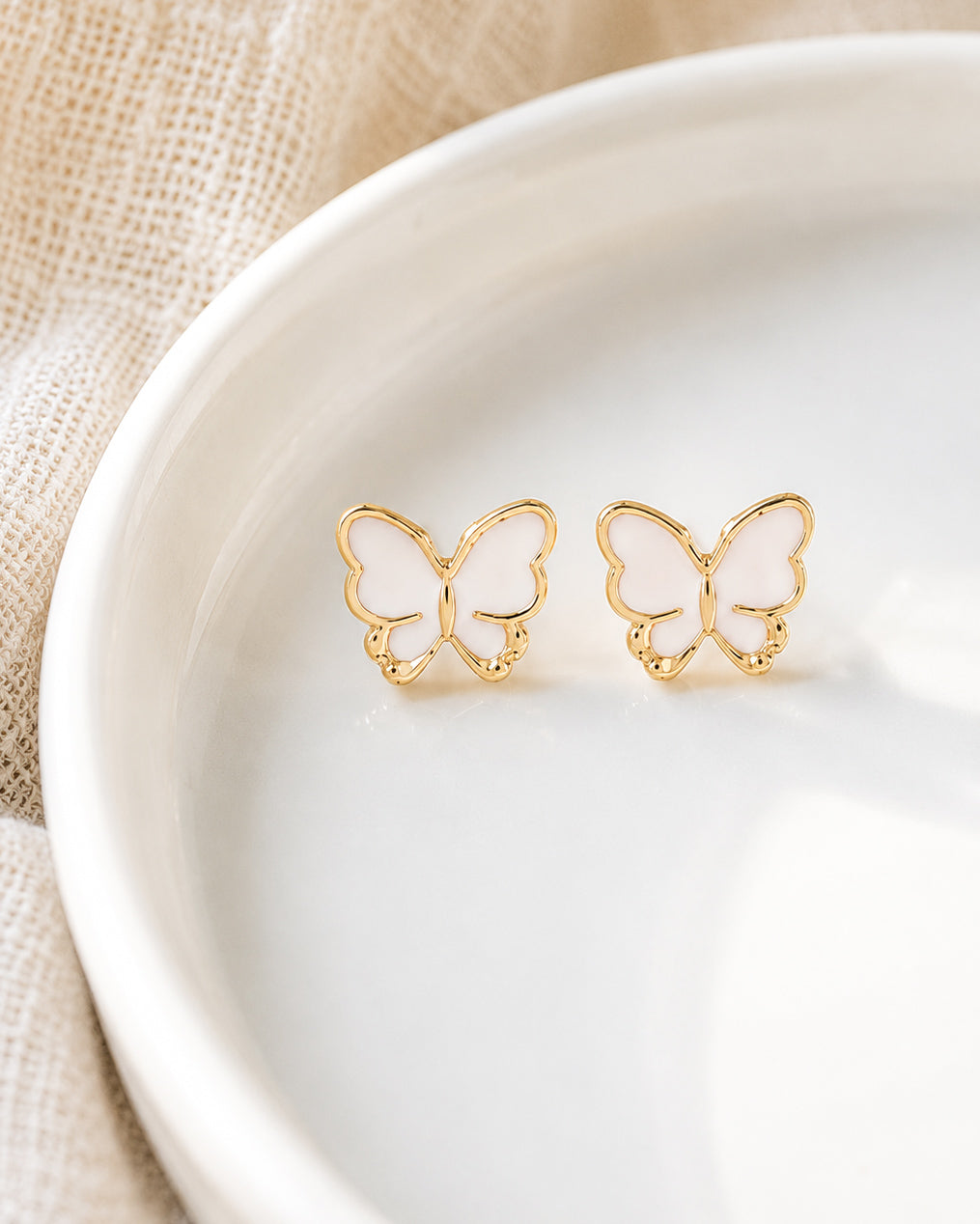 White and gold butterfly stud earrings resting in a white ceramic dish on soft neutral linen.