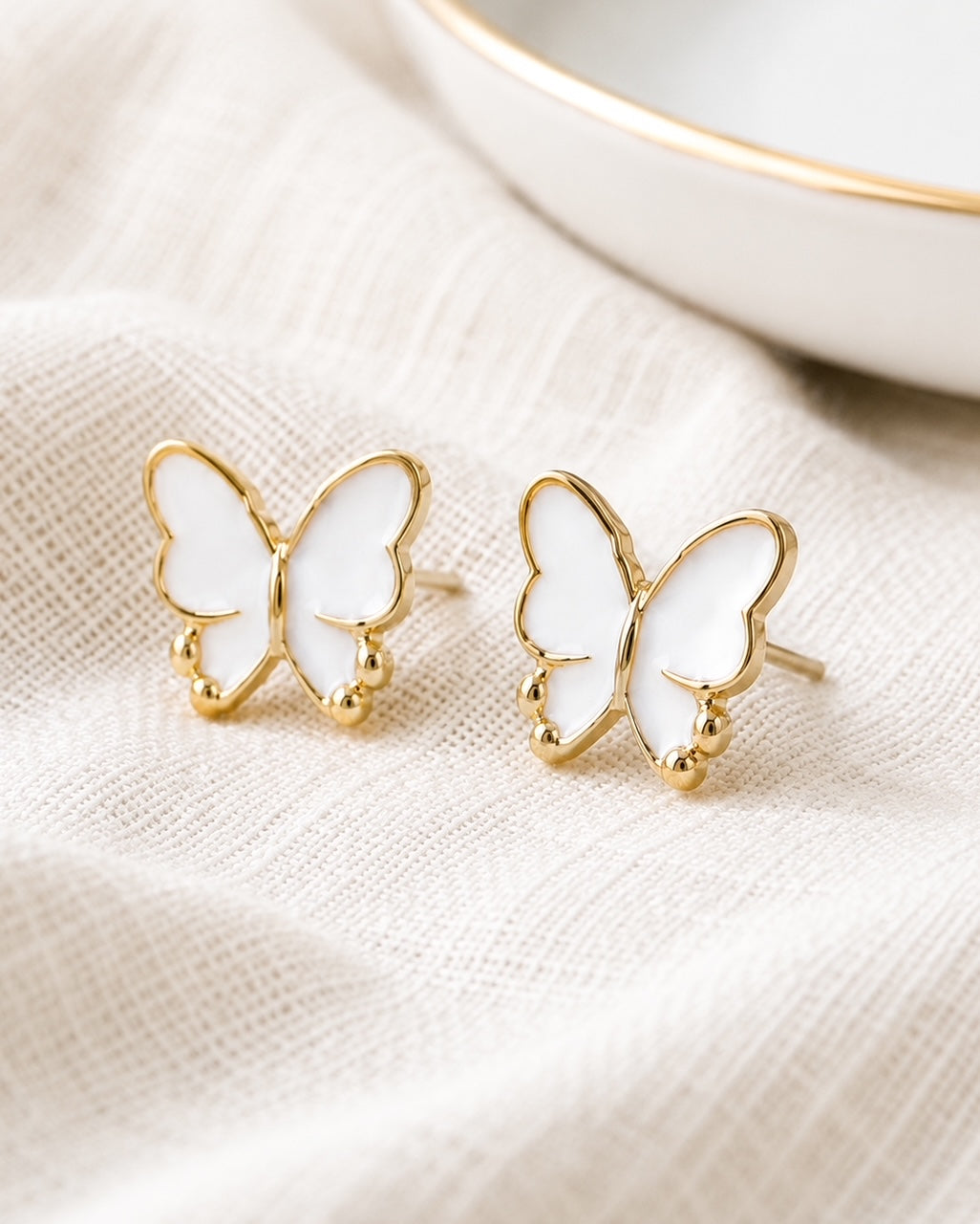 Delicate white butterfly stud earrings with gold-tone accents styled on soft neutral linen.