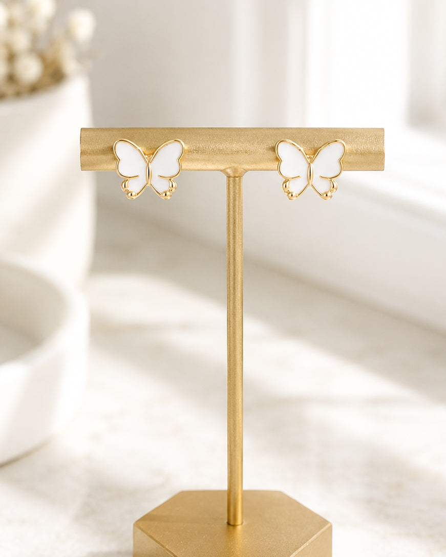 White and gold butterfly stud earrings displayed on a matte gold jewelry stand.