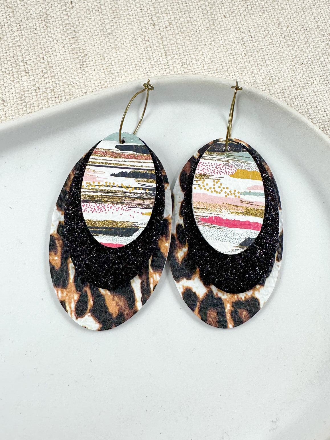 Layered oval leather earrings with leopard print base, black textured middle layer, and abstract patterned top layer on gold hoops displayed on a white dish