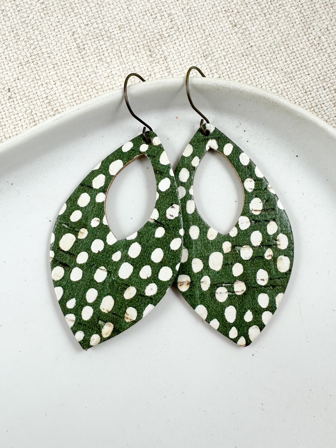 Flat lay of teardrop-shaped leather earrings with olive green background and white polka dots, displayed on a white ceramic dish.